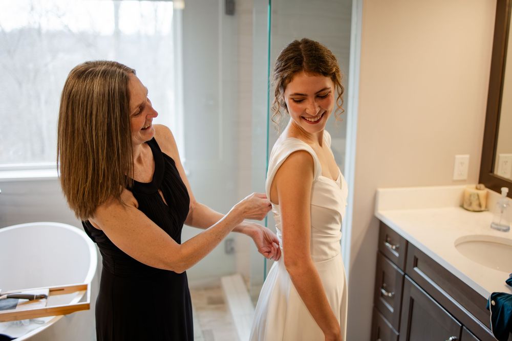A bridesmaid in black dress helps smiling bride button her white wedding gown in an elegant bathroom.