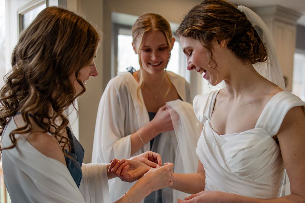 Bride in white gown getting ready with two bridesmaids in white robes, sharing a joyful moment.