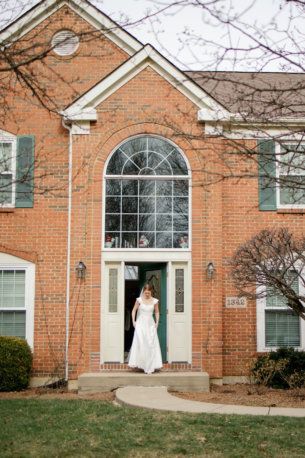 Bride in white wedding gown standing at the entrance of a brick colonial home with a green front door.
