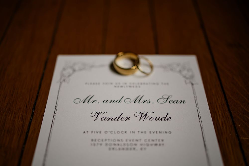 Wedding invitation for Mr. and Mrs. Sean Vander Woude with two gold rings on a wooden surface.