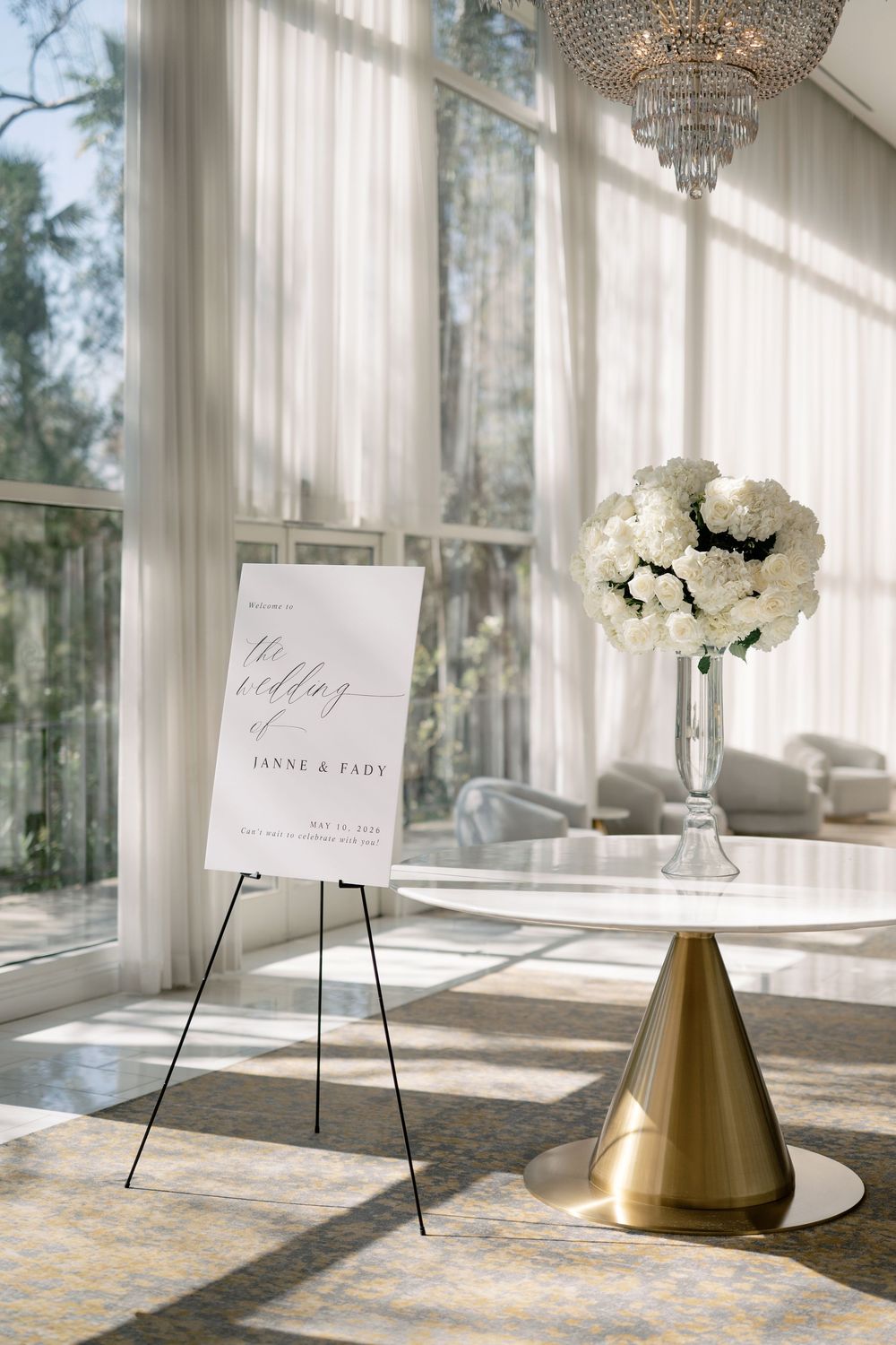 Elegant wedding welcome sign for Janny and Fady beside white floral arrangement in sunlit venue.