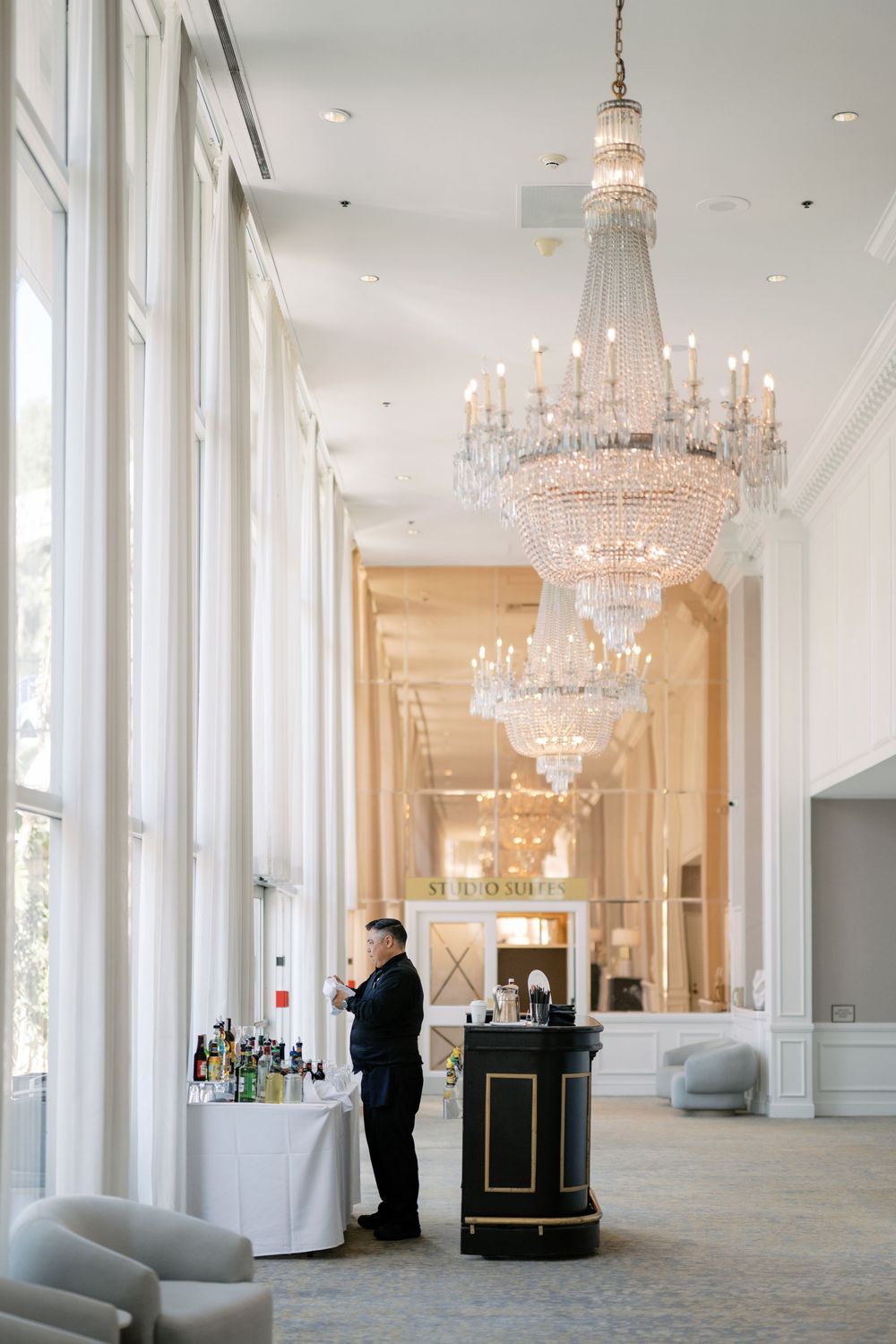 Elegant wedding venue hallway with grand crystal chandeliers and a bartender at a beverage station.