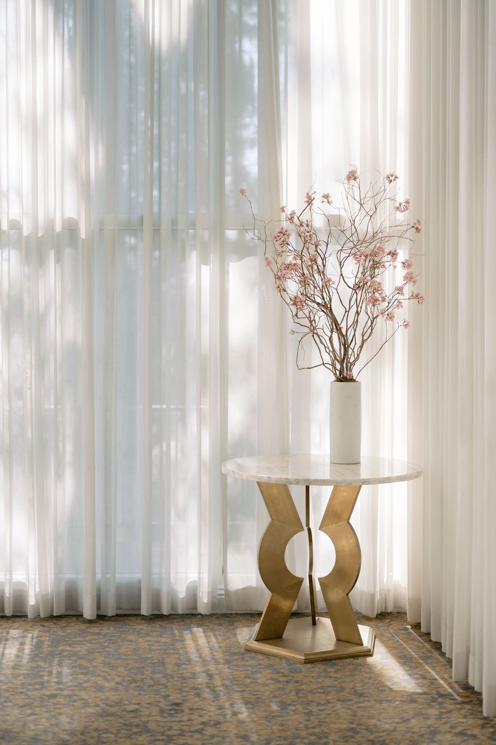 White vase with pink branches on gold marble table against sheer curtains in elegant venue.