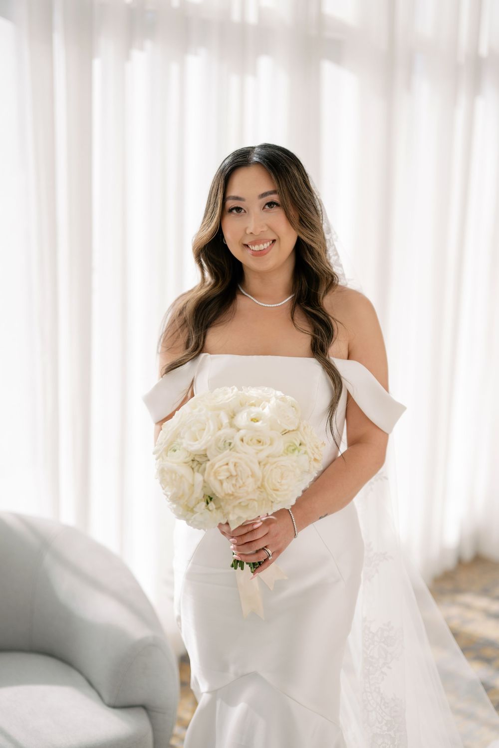 Smiling bride in off-shoulder white gown holding elegant white rose bouquet, standing by sheer curtains.