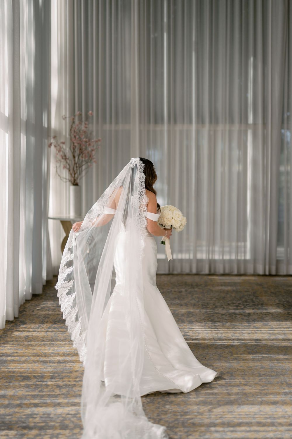 Elegant bride in a white fitted gown with a long lace-trimmed cathedral veil holding a bouquet by sheer curtains.