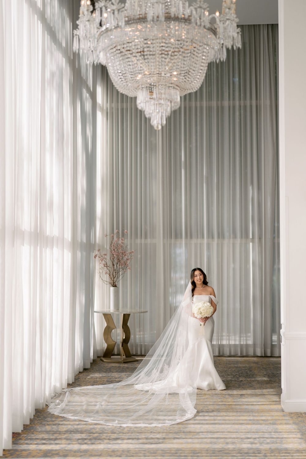 Bride in white mermaid gown with long veil stands beneath crystal chandelier in elegant venue.