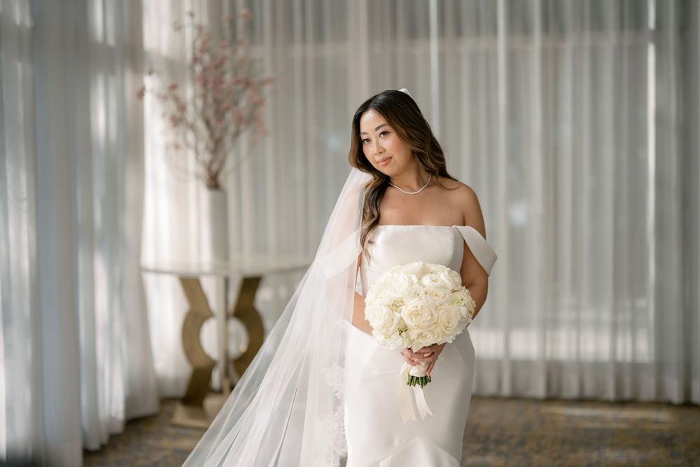 Elegant bride in off-shoulder white gown holding white bouquet with flowing veil.