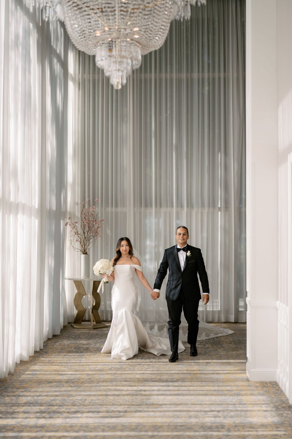 Bride in white mermaid gown and groom in tuxedo walk hand-in-hand under crystal chandeliers.