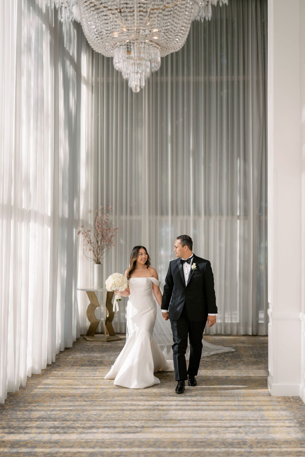 Bride and groom walk hand-in-hand down an elegant chandelier-lit hallway in wedding attire.