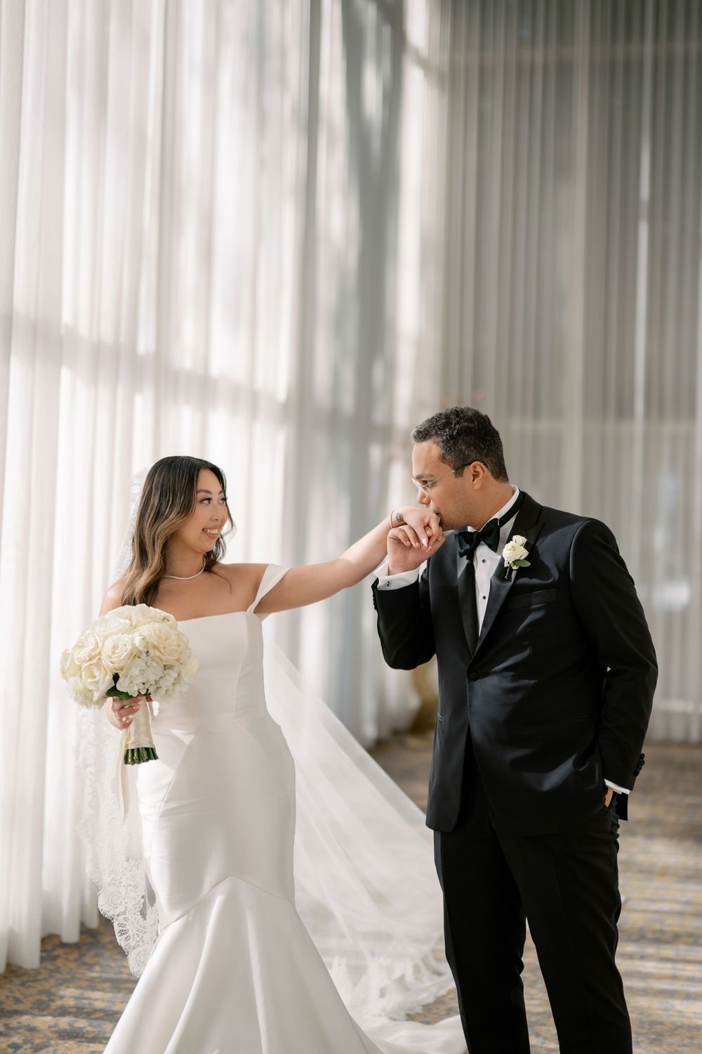 Groom in black tuxedo kissing bride's hand as she holds a white bouquet by sheer curtains.