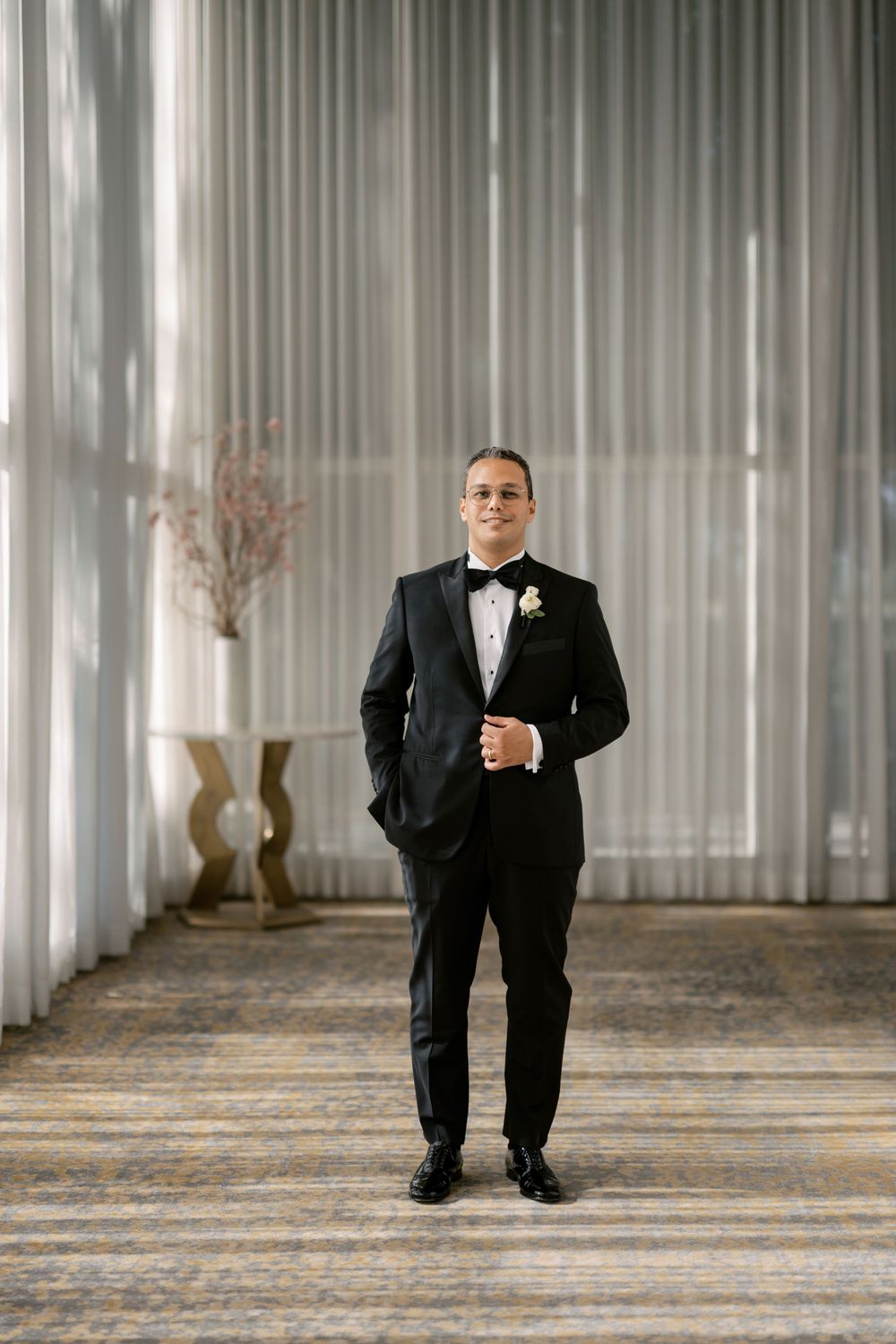 Groom in classic black tuxedo with white boutonniere poses in elegant hotel corridor.