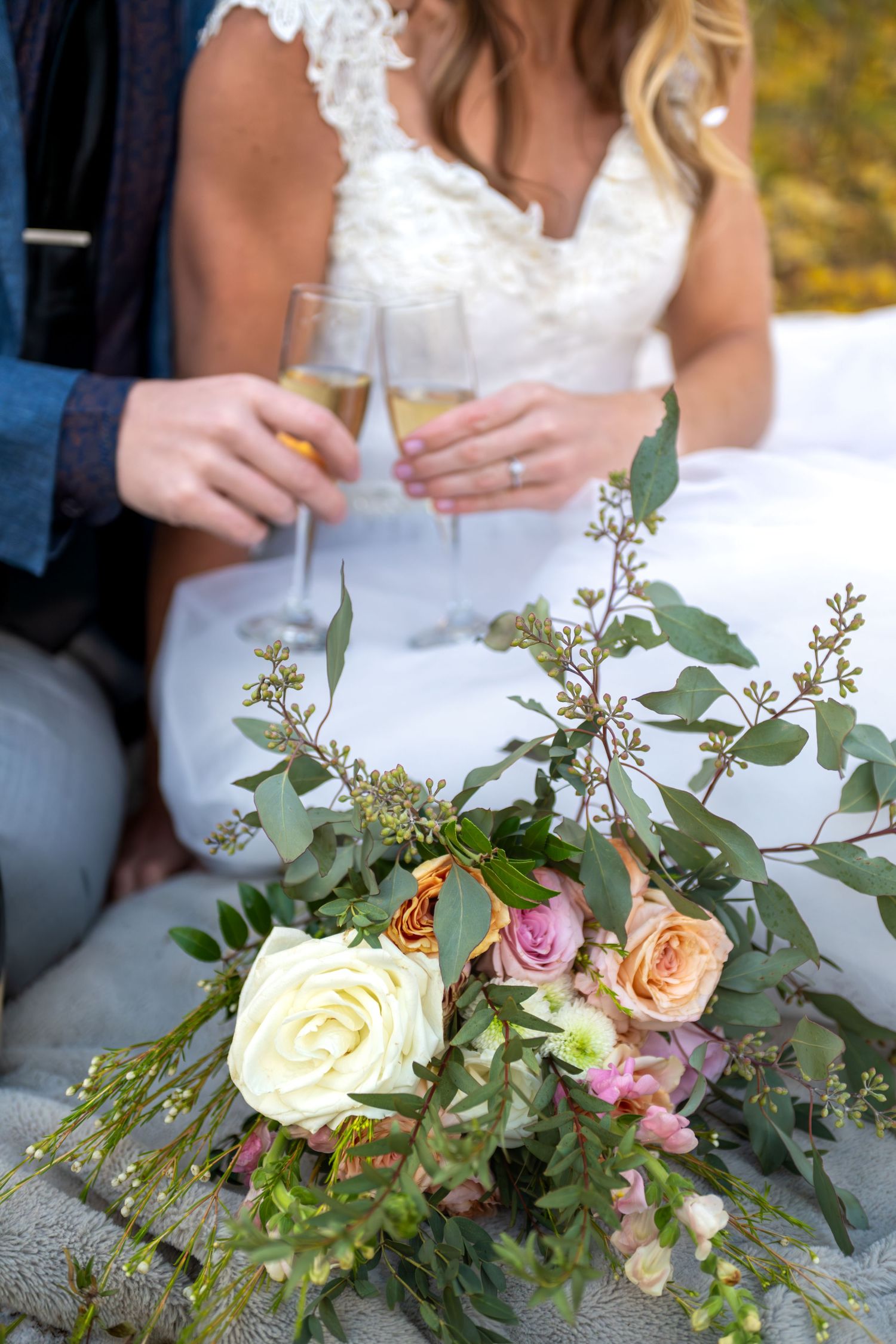 A close up image of a wedding bouquet of roses and eucalyptus in foreground with bride and groom toasting champagne glasses behind.