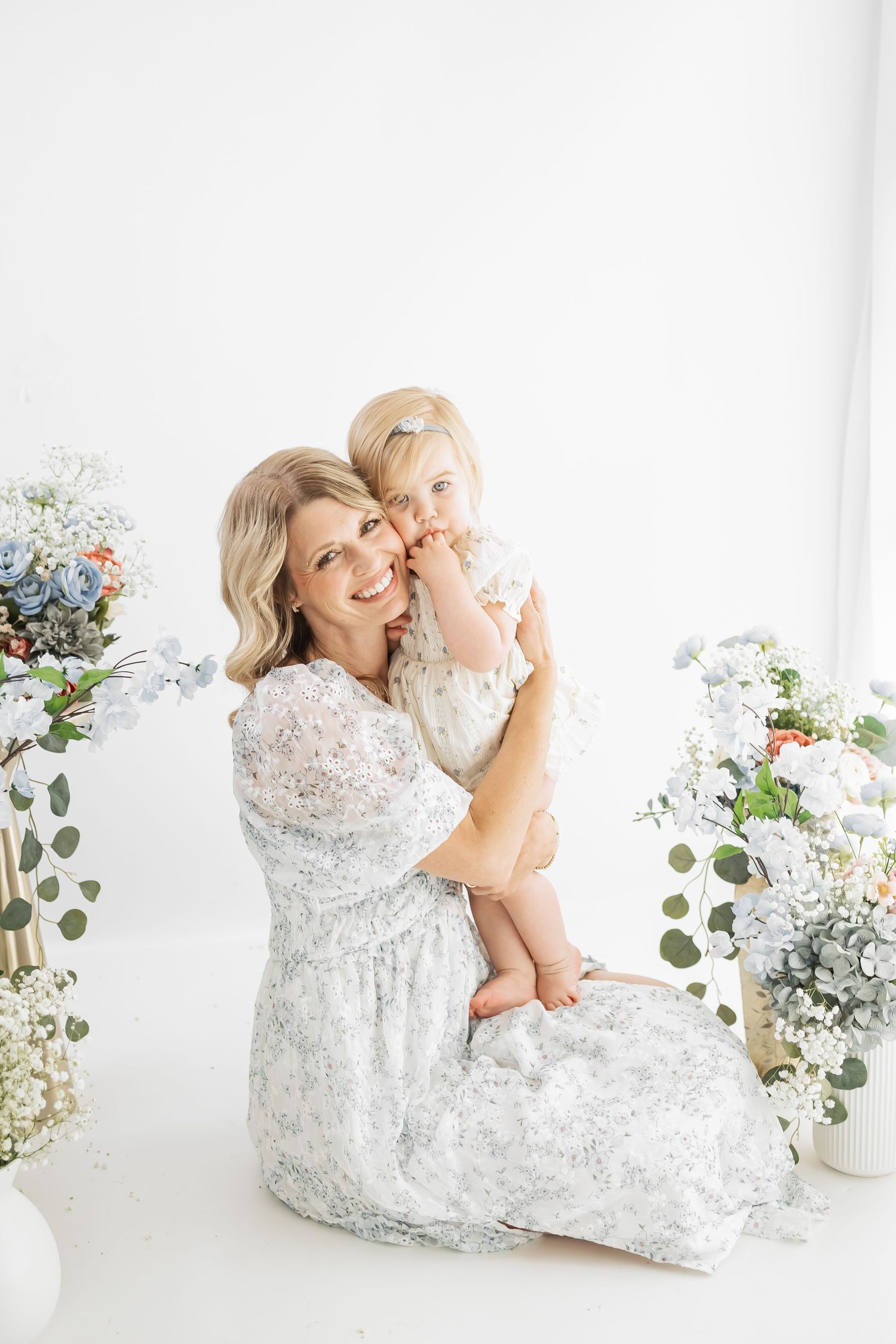 A mother in a floral dress holds her toddler daughter, surrounded by blue and white flowers in a bright studio.