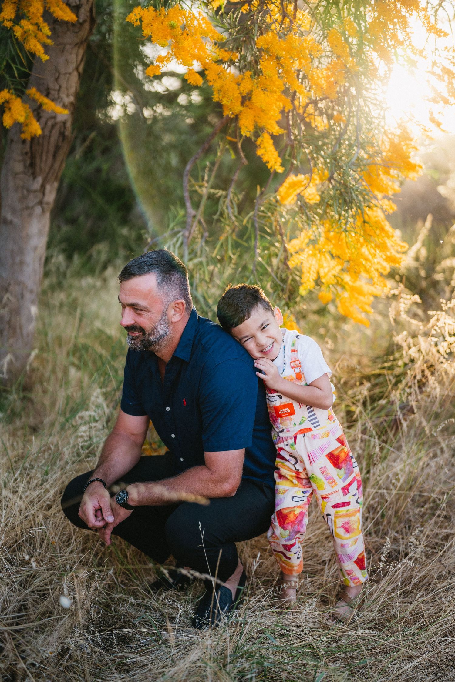 A man crouching outdoors as a young child hugs him from behind, surrounded by golden autumn foliage in warm sunlight.