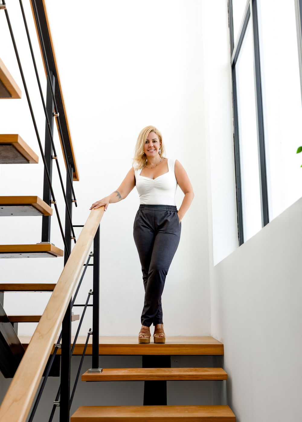 Blonde woman in white top and dark pants posing on modern wooden staircase with black metal railing.