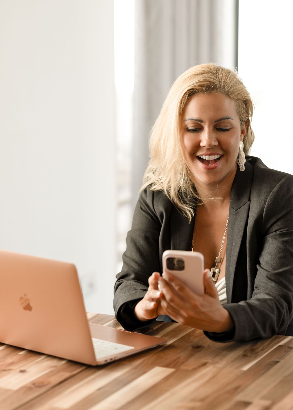 Smiling blonde businesswoman in blazer using smartphone at wooden desk with rose gold MacBook laptop.