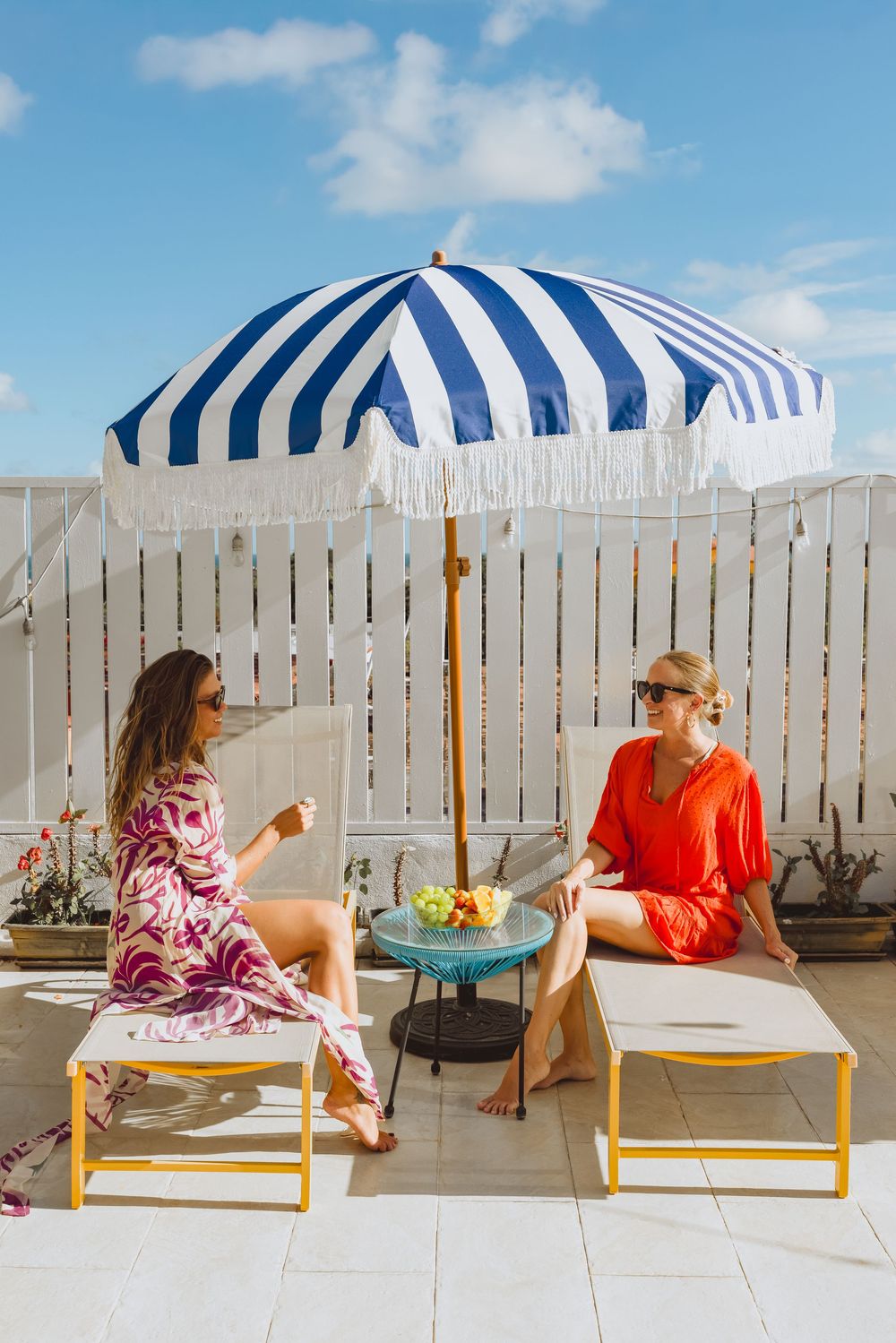 Two women in colorful summer outfits relax on yellow lounge chairs under a blue striped umbrella on a sunny patio.