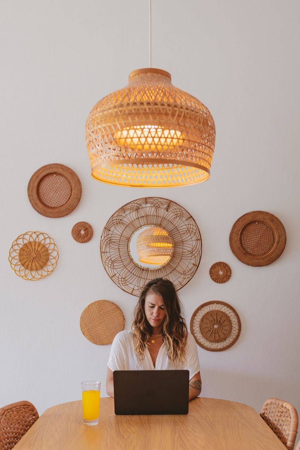 Woman working on laptop at wooden table under woven rattan pendant light with decorative wall baskets and orange juice.
