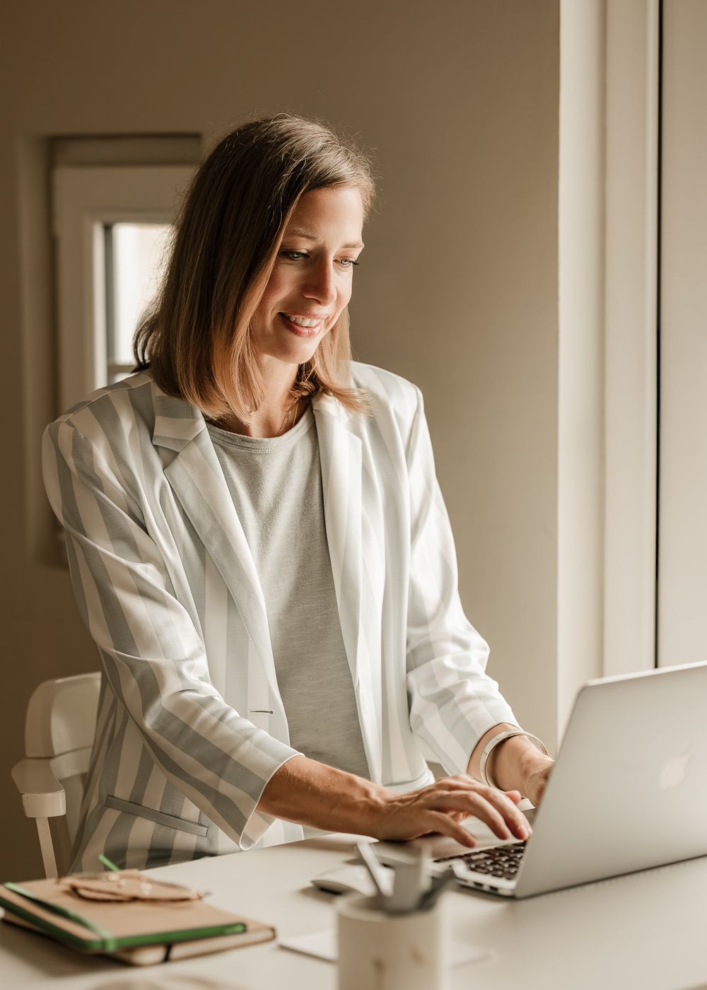 Woman in white blazer smiling while working on laptop in bright office setting.