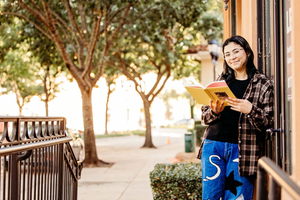 Young woman smiling while reading a yellow book outdoors, leaning against a wall on a sunny urban sidewalk.