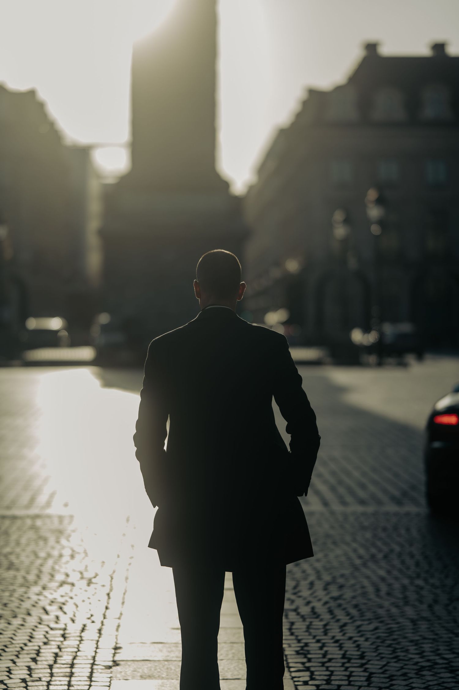 Un homme d'affaires en silhouette se tient sur une rue pavée au coucher du soleil avec des bâtiments urbains et un rétroéclairage doré.