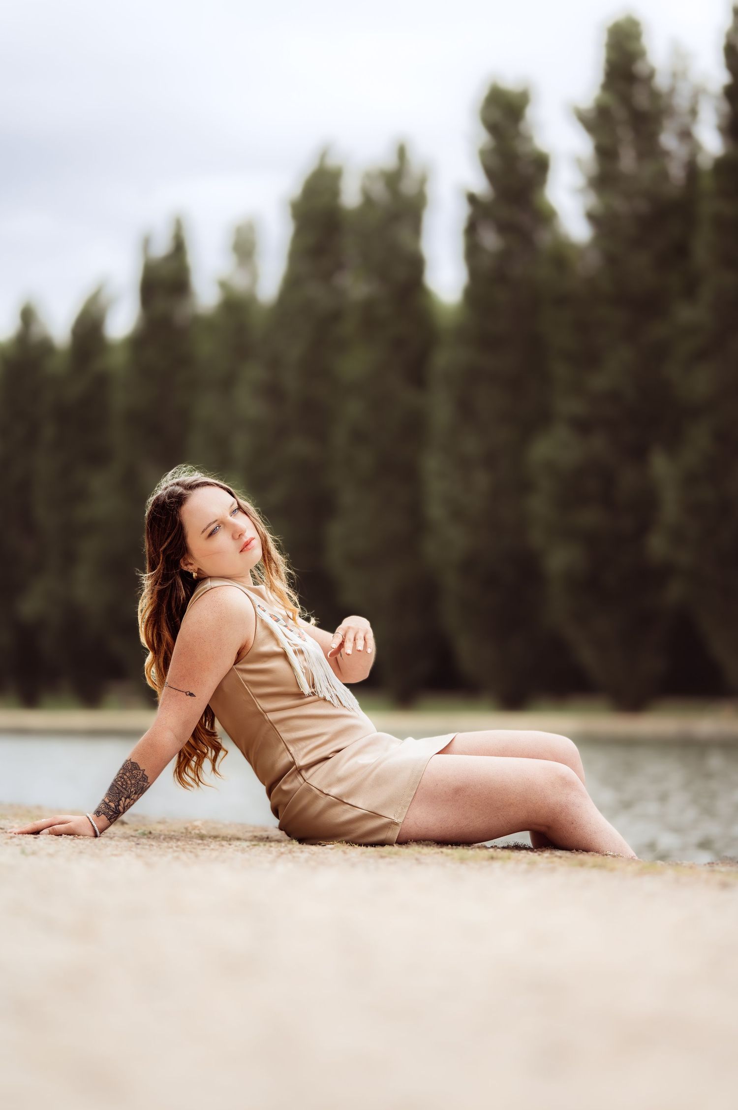 Femme en robe beige assise sur le rivage de sable avec de grands arbres à feuilles persistantes et un lac en arrière-plan.