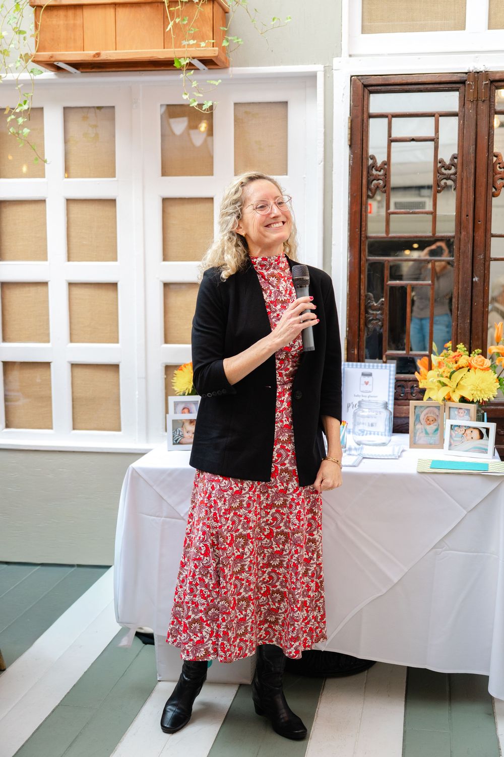 Woman in floral dress and black blazer holds microphone at baby shower event.