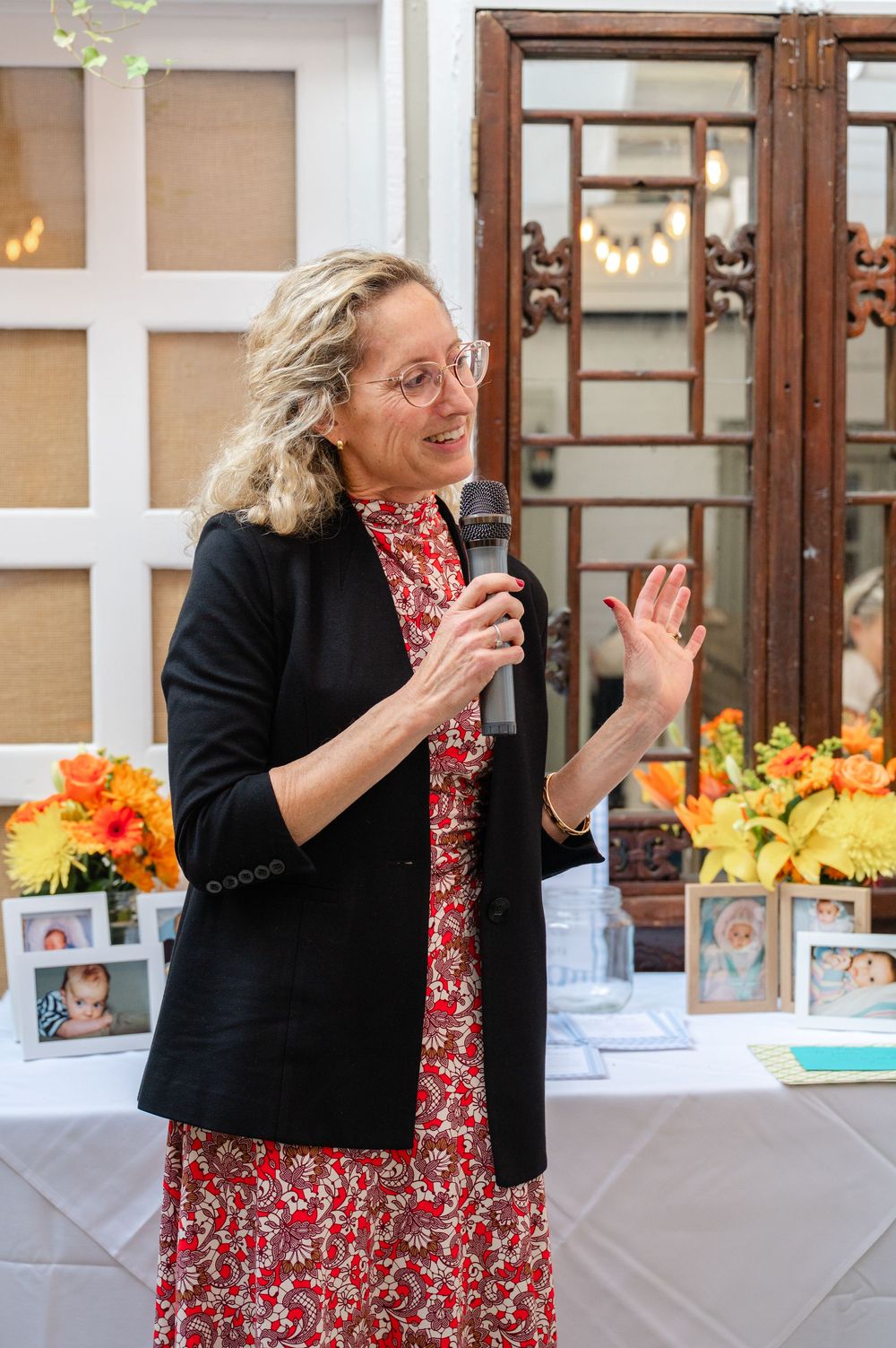 Woman in red floral dress and black blazer speaks into microphone at elegant indoor event with flowers.