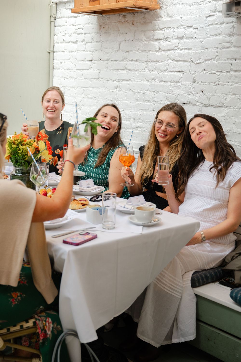 Women laughing and raising cocktails at a white-tablecloth restaurant table with brick wall backdrop.
