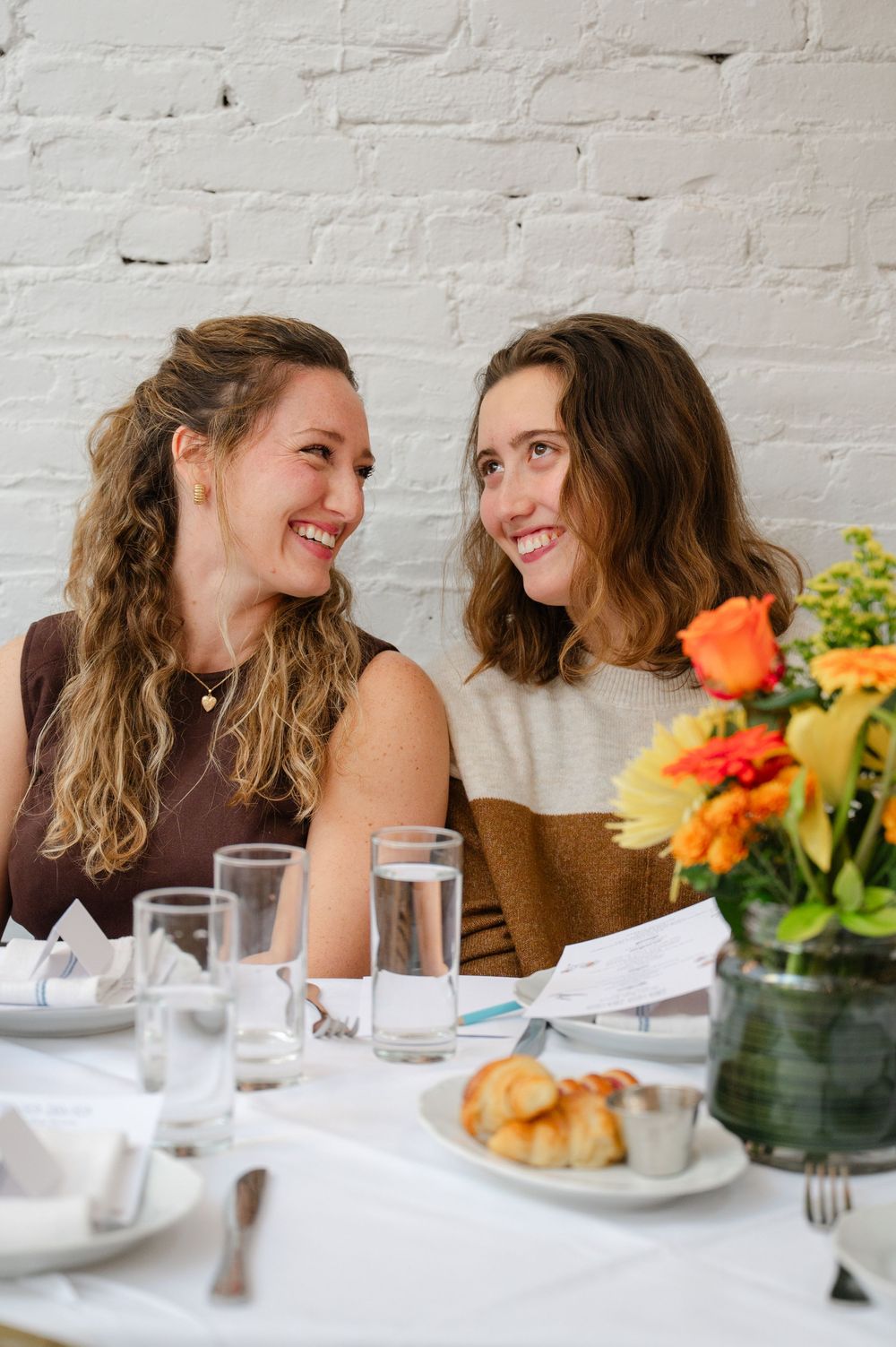 Two smiling women sharing a joyful moment at a dining table with colorful flowers and water glasses.