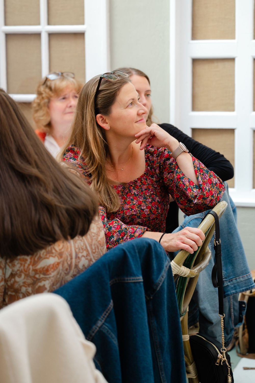 Woman in floral top sits thoughtfully at an indoor gathering, resting chin on hand while listening attentively.