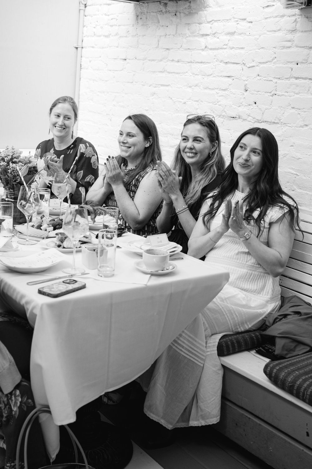 Four women smile and clap together at a restaurant table during a celebration event.