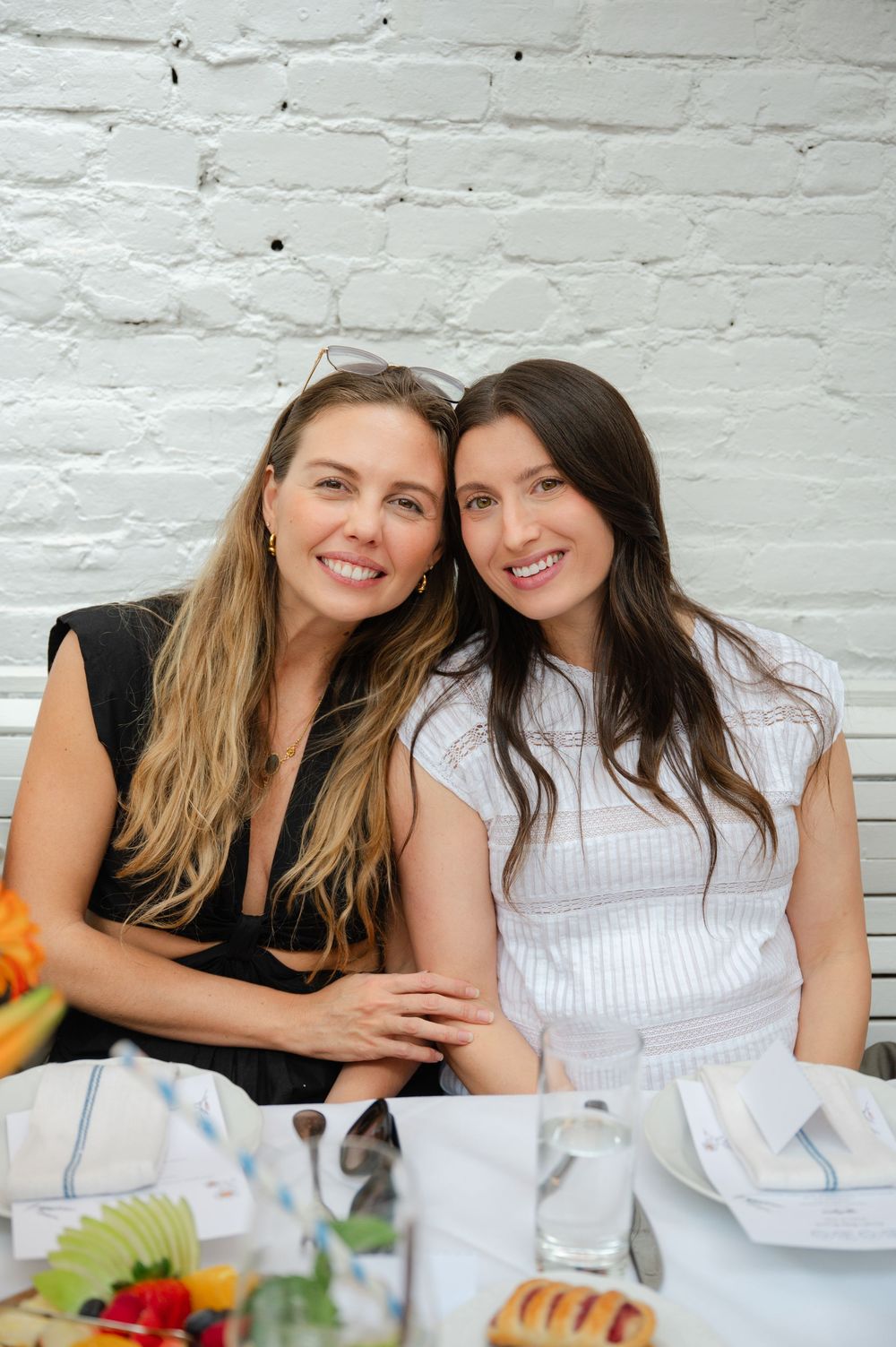 Two smiling women posing together at a brunch table with colorful fruit.