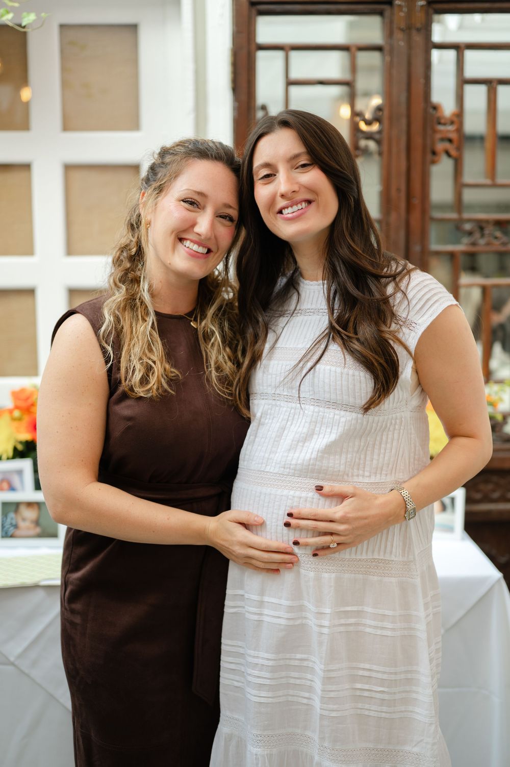 Two women smiling at a baby shower, one in white dress cradling baby bump.