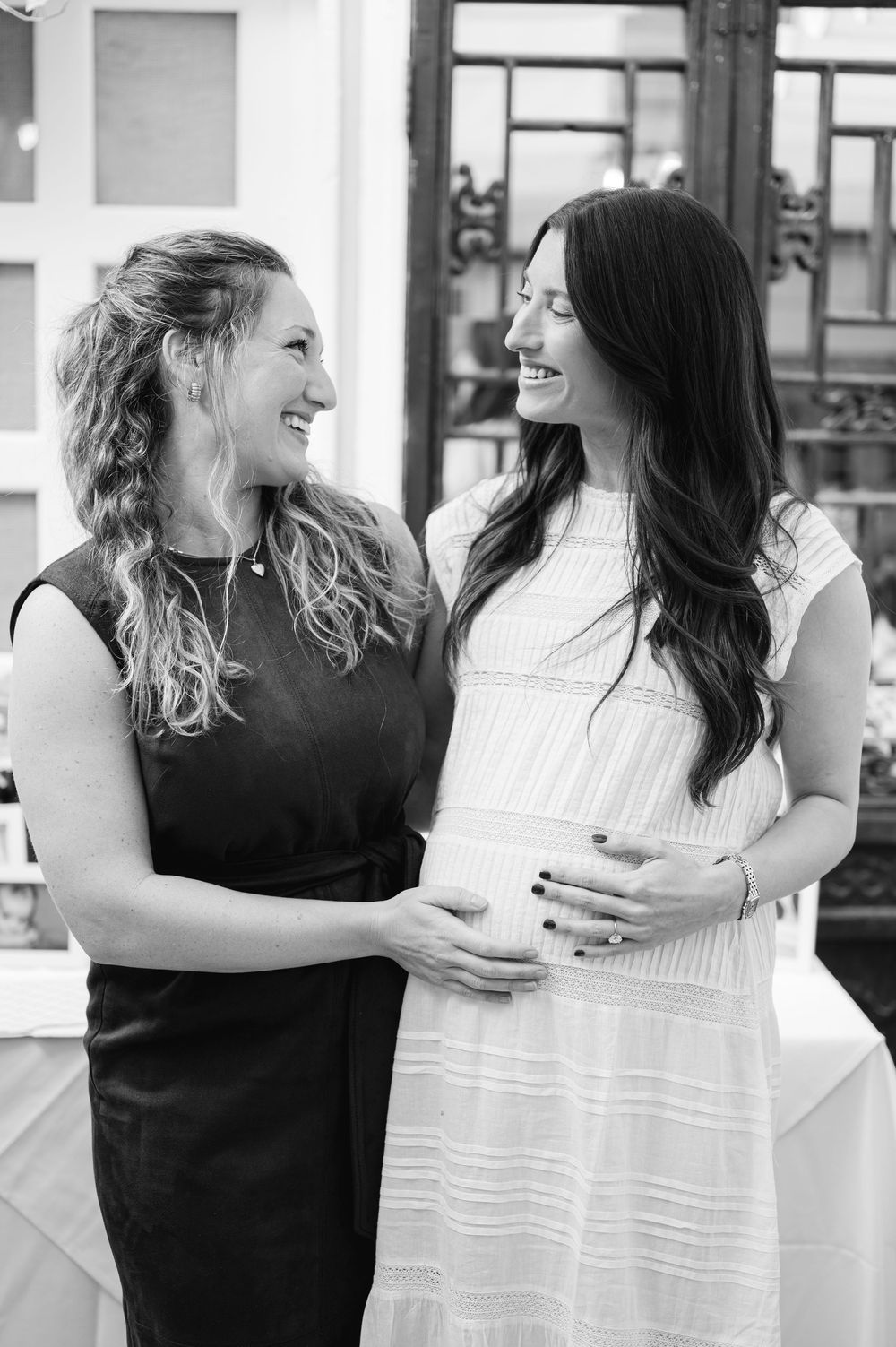 Two women smiling at each other, one in black dress touching pregnant belly of woman in white dress, black and white photo.