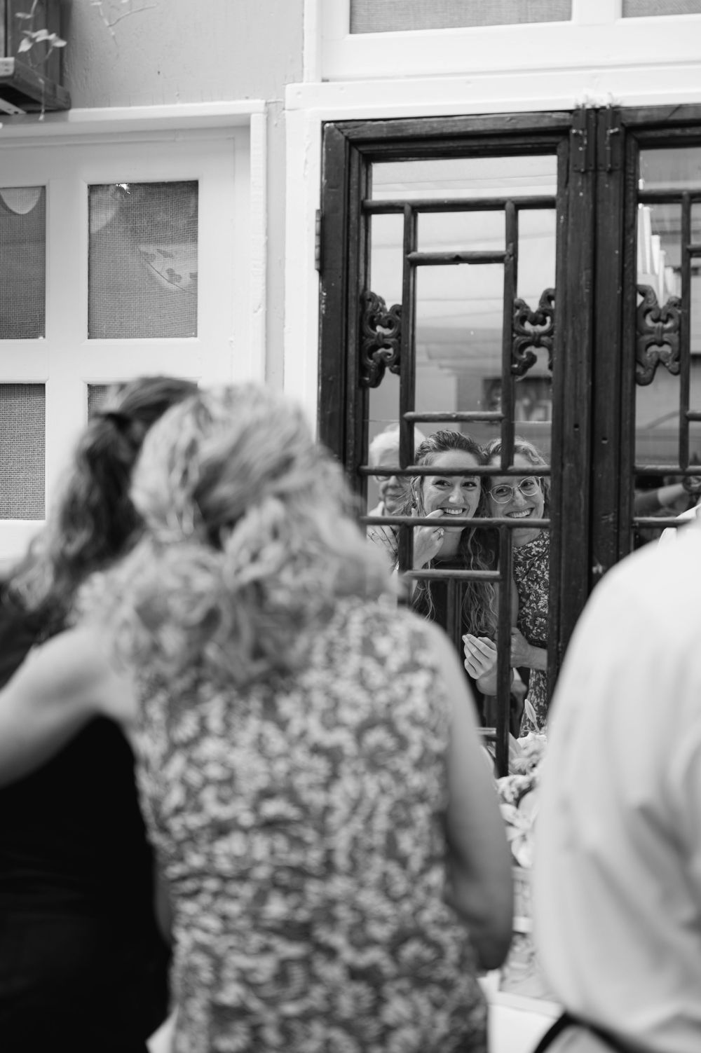Woman peers through ornate glass doors at a gathering in black and white photo.
