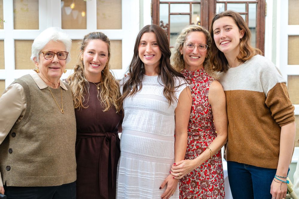 Five women smiling and posing together indoors at a social gathering, dressed in elegant casual attire.