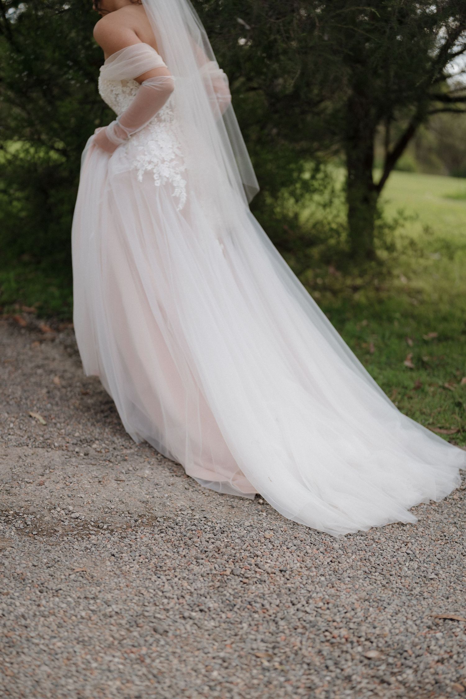 Bride in a flowing white tulle ball gown with lace bodice and sheer gloves, standing on a gravel path outdoors.