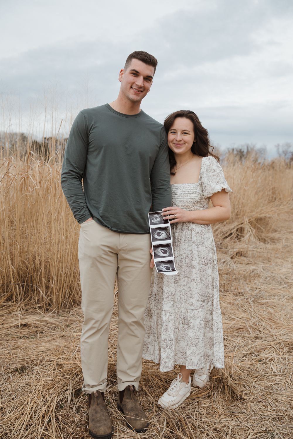 A couple stands in a golden field holding ultrasound photos, announcing their pregnancy outdoors.