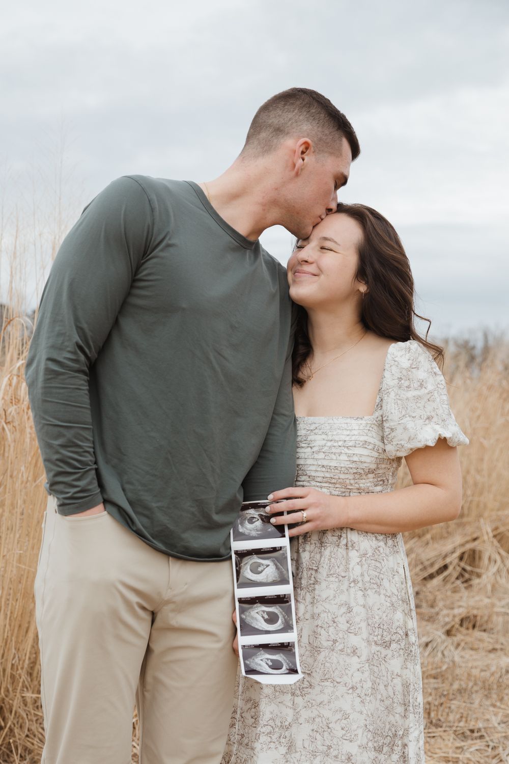 Happy couple holding ultrasound photos outdoors in a field, sharing a tender kiss during pregnancy announcement.
