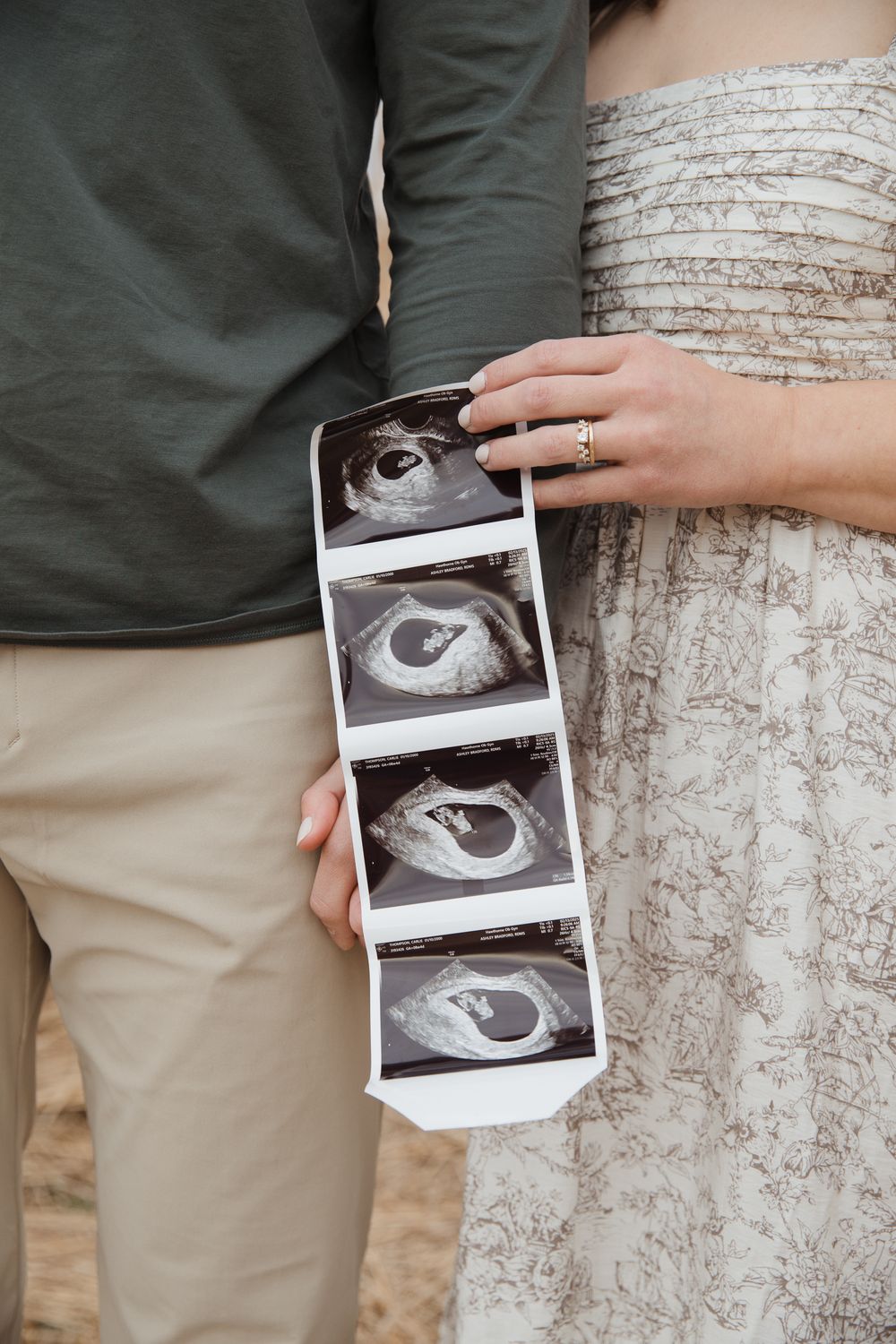 Couple holding ultrasound sonogram photos, woman wearing floral dress and gold ring, pregnancy announcement photo.