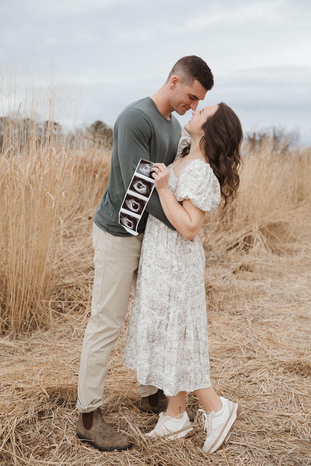 Couple embracing in a golden field holding ultrasound photos, announcing pregnancy outdoors.