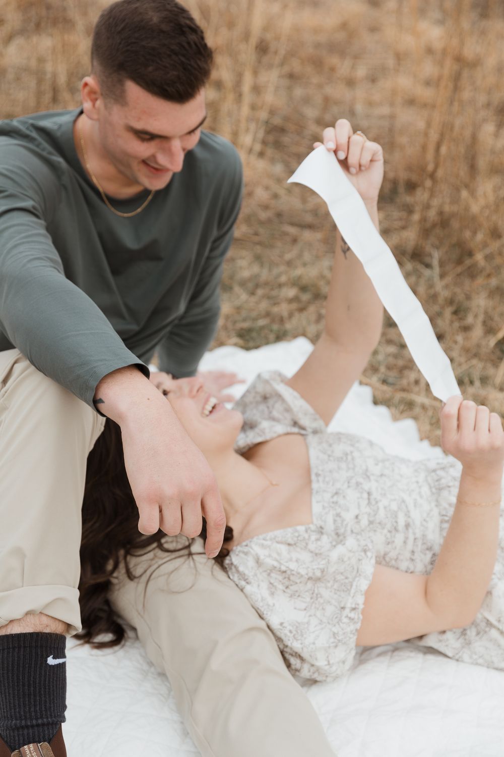 Smiling couple sitting in a field, woman lying down holding ultrasound strip during pregnancy announcement.