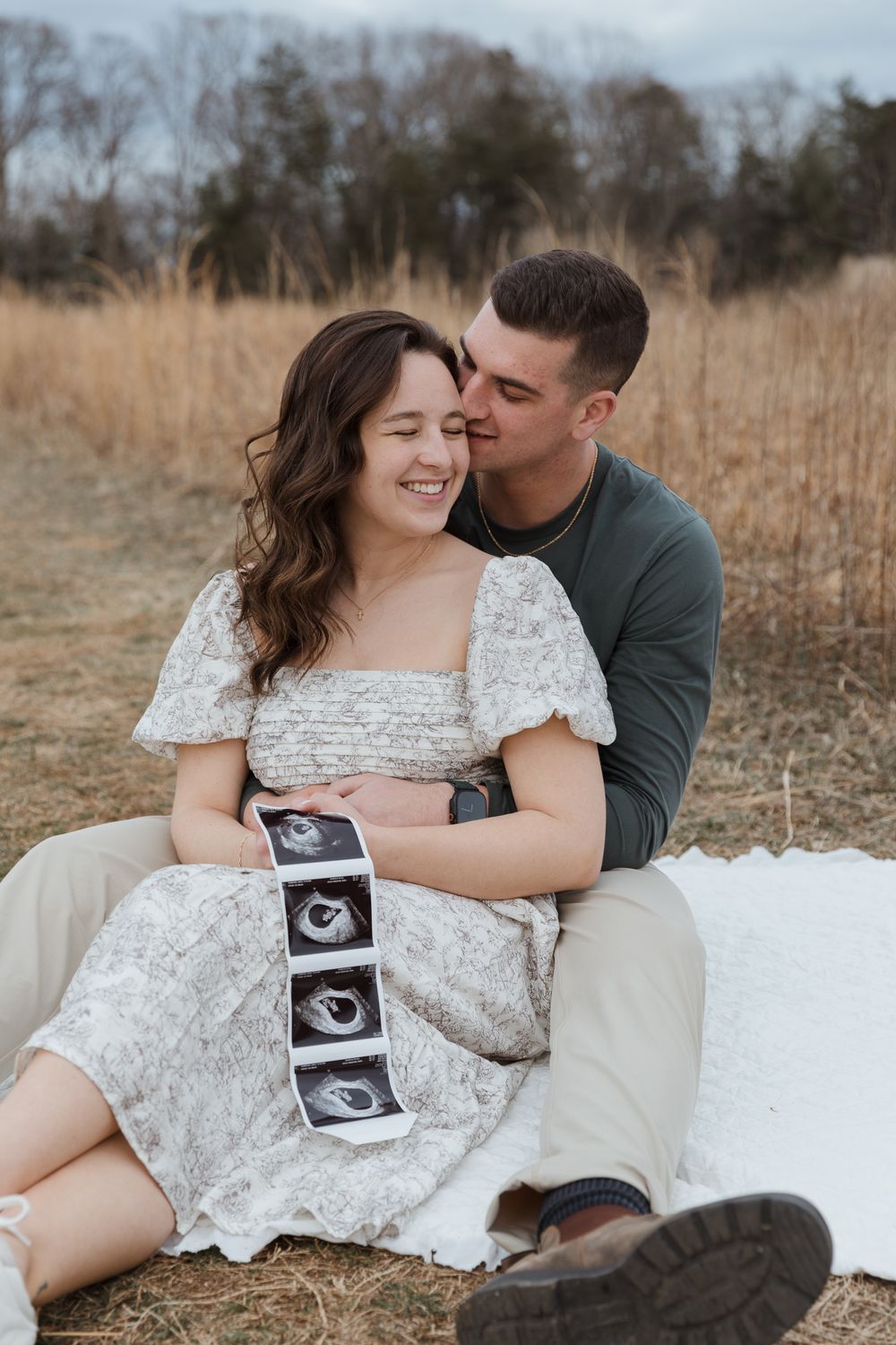Happy couple sitting in a field holding ultrasound photos, celebrating pregnancy announcement outdoors.