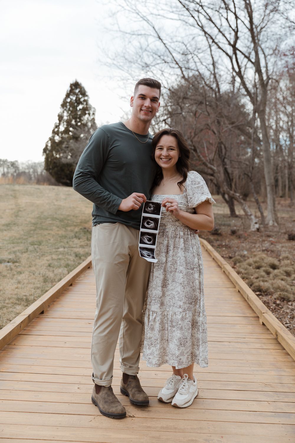 Happy couple holding ultrasound photos on a wooden boardwalk, announcing their pregnancy outdoors.