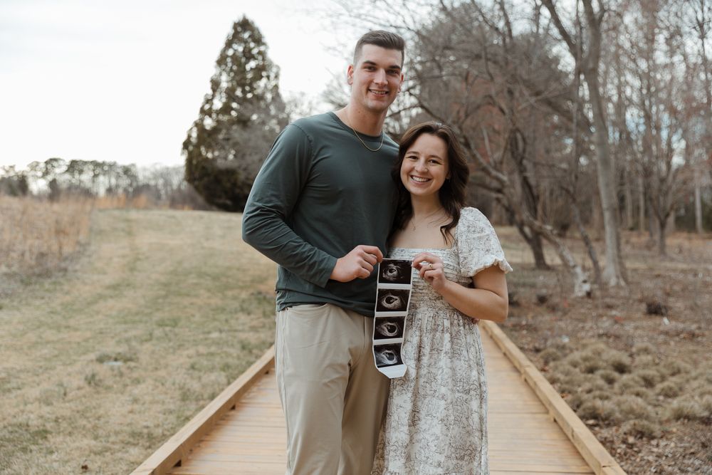 Happy couple standing on a wooden boardwalk holding ultrasound photos announcing their pregnancy outdoors.