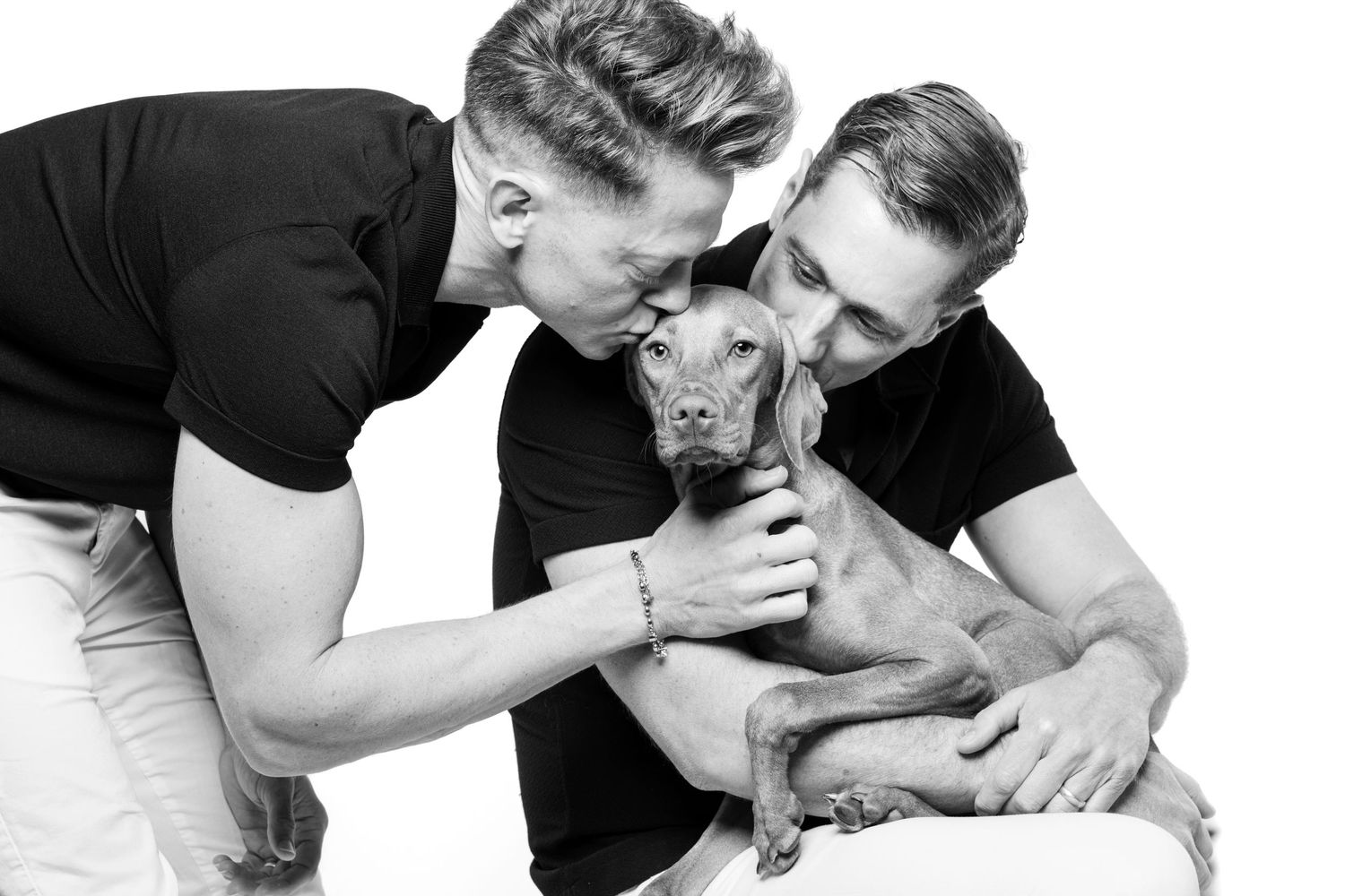Two men in black shirts lovingly kiss a small brown Vizsla in a black and white studio portrait.
