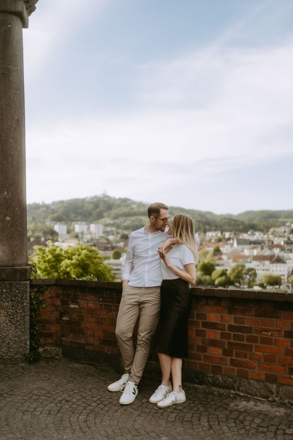 Couple in white shirts sharing a romantic moment on a brick terrace overlooking a European city skyline.