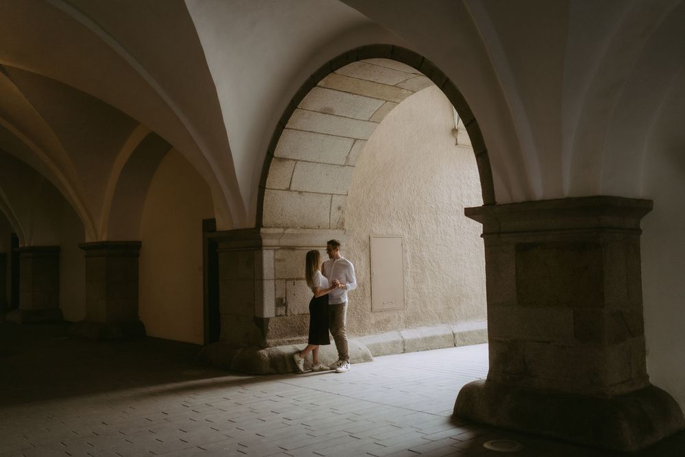Couple embracing romantically under a stone archway in an ancient European building with vaulted ceilings.