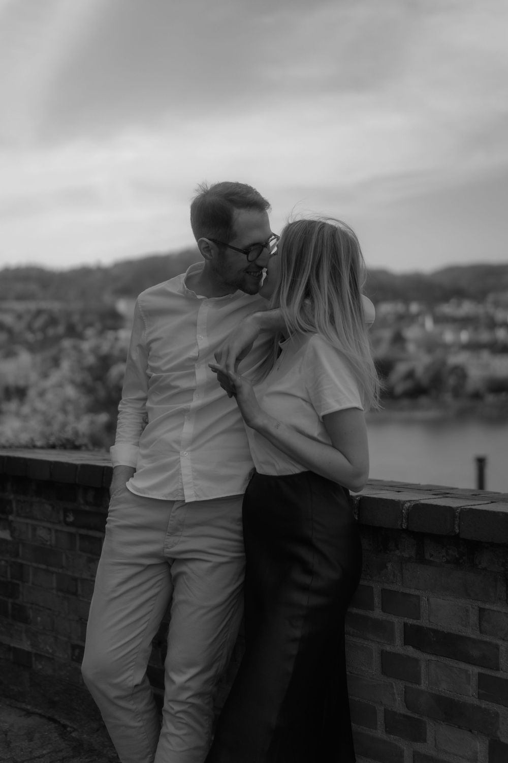 Black and white photo of a couple sharing a kiss on a brick wall overlook with a river view.
